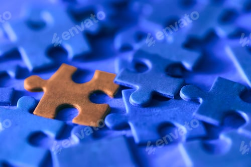 Preview: Single Orange Puzzle Piece Resting Among Scattered Blue Puzzle Pieces On Textured Surface Closeup
