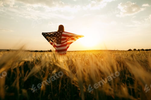 Preview: Young Woman with the American flag running in a wheat field at sunset. 4th of July.