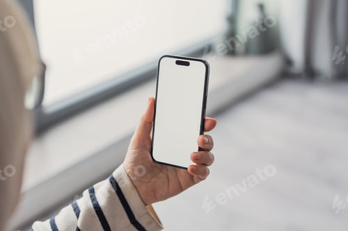 Preview: Person holding smartphone with blank screen near window in modern home interior for mockup
