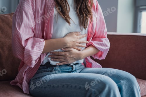 Preview: Young woman sitting on couch holding her stomach in pain, experiencing abdominal cramps and menstrua