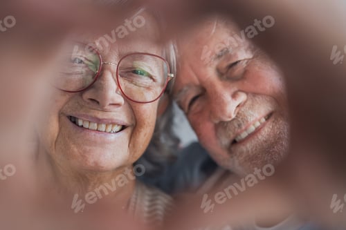 Preview: A happy retired couple poses indoors making a heart gesture with their fingers, smiling at the camer