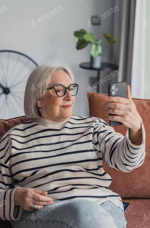 Preview: Senior woman taking a relaxed selfie on a smartphone while seated on a cozy living room couch
