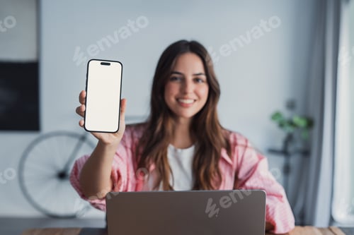 Preview: Smiling woman showing smartphone screen while working on laptop in bright home office