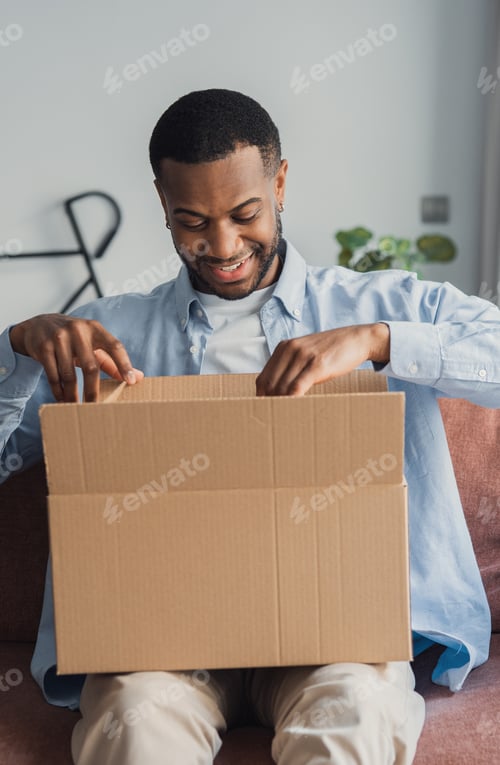 Preview: Smiling man happily unboxing a delivery package at home on a comfortable couch