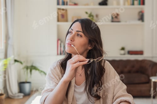 Preview: Thoughtful woman holding glasses and looking out window while working or reflecting at home