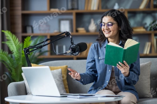 Preview: Smiling young Indian woman sitting on sofa at home, holding a book, reading and talking on laptop ca
