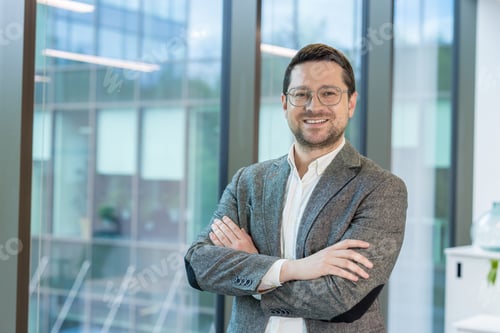 Preview: Portrait of a smiling young businessman standing in the office by the window in a suit and glasses,