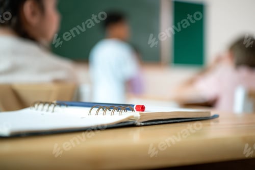 Preview: Notebook and pencil on desk with blurred classroom background