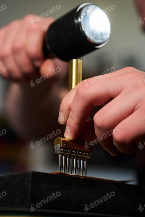 Preview: Close up of artisan hands using a heavy mallet and a brass stitching punch to make holes in leather.