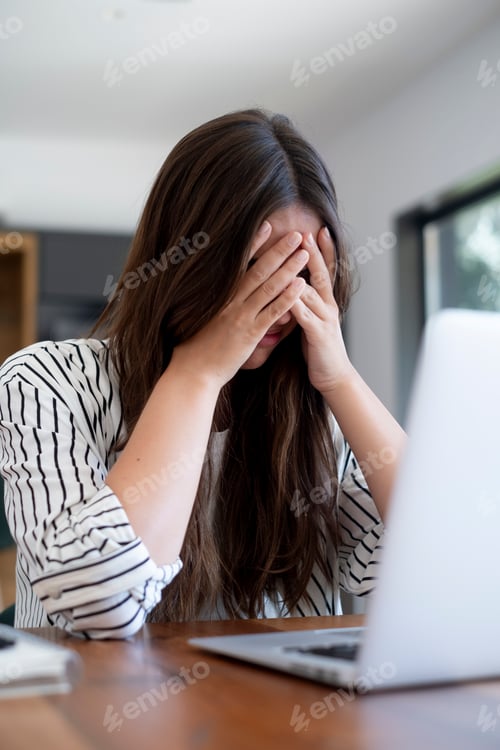 Preview: Stressed young woman covering her face while working on a laptop in a home office