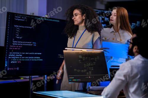 Preview: Black female IT analyst with curly hair works at a laptop while a red-haired female professional in