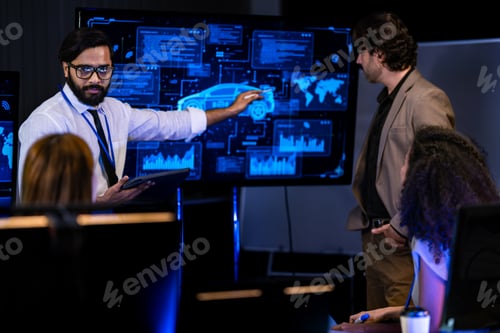 Preview: Tech team leader in a tan blazer addresses seated colleagues in a dark briefing room lit by blue AI