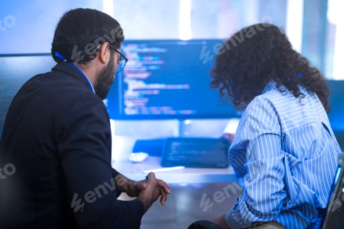 Preview: A male team lead leaning over a seated female developer pointing at a line of code on the screen, to