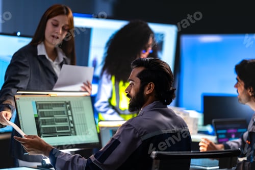 Preview: Bearded male engineer examines a document held up beside a glowing monitor in a dark engineering off