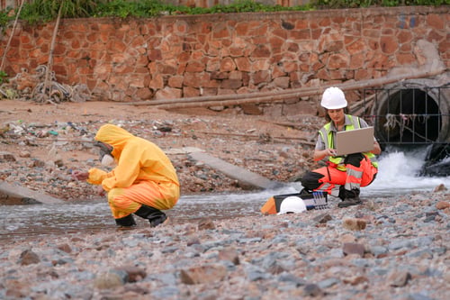 Preview: Environmental field team in protective gear analyzes water samples on a polluted beach. One holds a