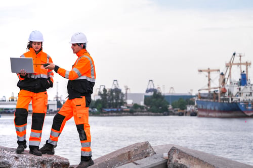 Preview: Two diverse engineers in safety gear coordinate a project at a waterside construction site. A female