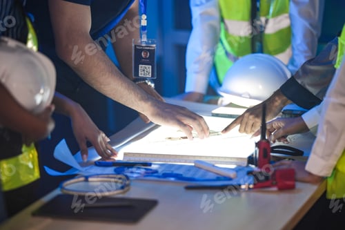 Preview: A close up of an engineering team collaborating at night. Hands point at a glowing blueprint on a li