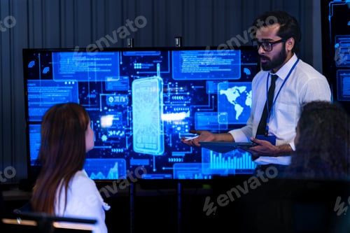 Preview: Male presenter in a tan blazer addresses seated tech colleagues in a dark briefing room with glowing