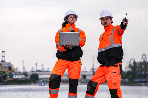 Preview: Diverse industrial engineers in orange safety uniforms and hard hats collaborating with a laptop at