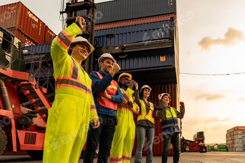 Preview: Happy diverse logistics team celebrating success by raising fists together at a container port. Succ