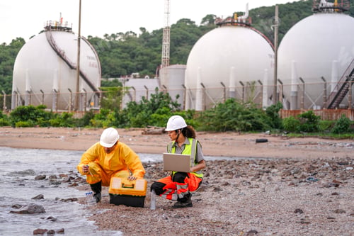 Preview: Two engineers in safety gear collect water samples and input data on a laptop near coastal chemical