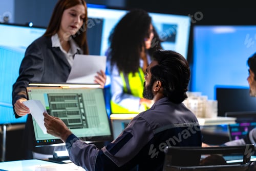 Preview: South Asian engineer reviews documents near a lit CAD monitor while a red-haired female professional