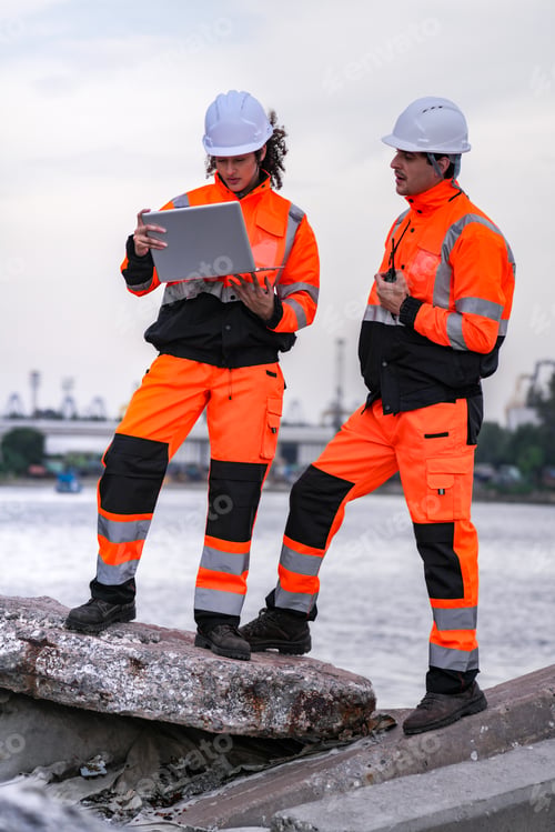 Preview: Two diverse engineers in safety gear coordinate a project at a waterside construction site. A female