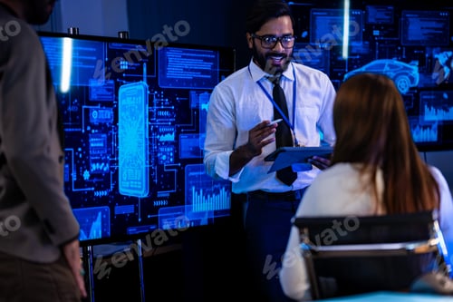Preview: Male presenter in a tan blazer addresses the team from the front of a dark briefing room with glowin