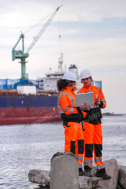 Preview: Two diverse engineers in safety gear coordinate a project at a waterside construction site. A female
