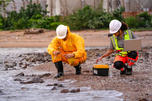 Preview: Two environmental engineers in safety gear work at a shoreline near gas storage tanks. One collects