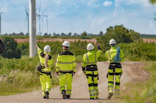 Preview: group of engineers in high visibility gear stands at a wind farm, observing several wind turbines. T