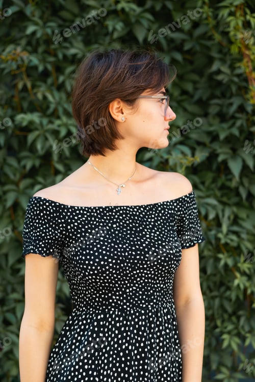 Preview: A woman stands facing to the side in a dark dress with a pattern of spots, surrounded by lush green