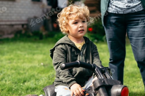 Preview: A young child with curly hair sits on a toy vehicle in a grassy area, looking pensive as an adult st