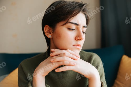 Preview: A young woman sits indoors, lost in thought with her hands clasped near her face. She has short hair