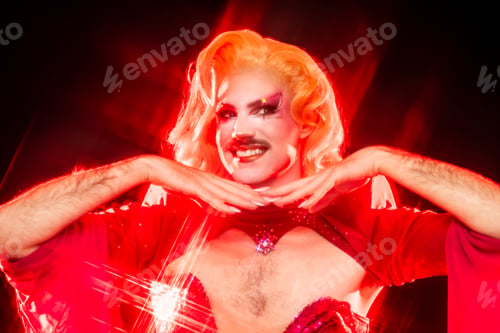 Preview: Smiling Drag Queen in Red Outfit Poses With Hands Near Face in Studio Setting