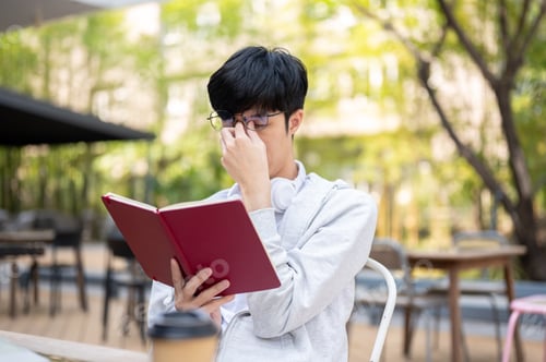 Preview: Glasses man rubbing eyes feeling tired or sleepy while reading book sitting at table in garden cafe.