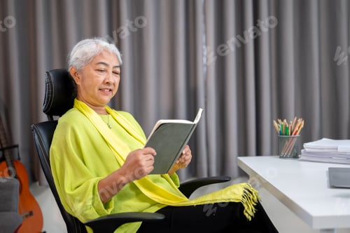 Preview: Happy elderly old asian woman reading a book while sitting leaning on chair in a living room at home