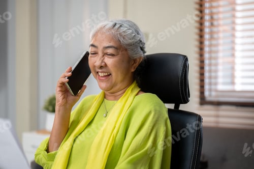 Preview: Laughing elderly senior old asian woman holding paper talking on phone call at table in home office.