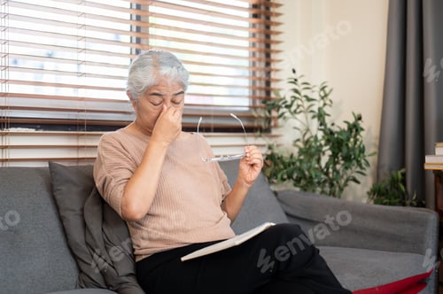 Preview: Old asian woman grandma taking off glasses massaging nose as sitting on sofa in living room at home.