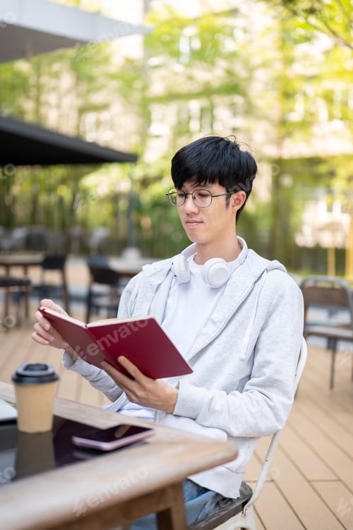 Preview: Asian man wearing glasses reading a book over phone and coffee while sitting at table in garden cafe