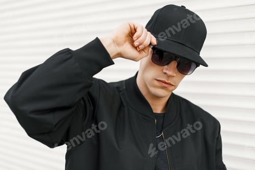 Preview: Handsome man with black fashionable glasses and a black baseball cap posing near a white wall