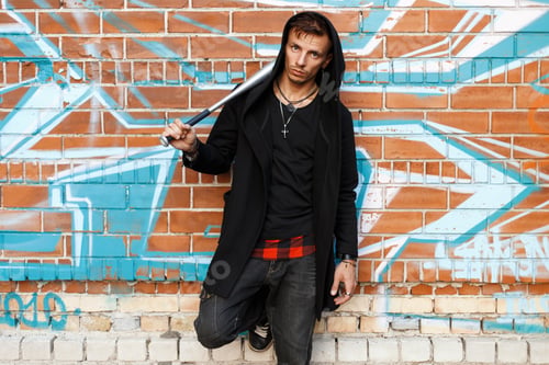 Preview: Stylish handsome man with a bat is posing near brick wall with graffiti.