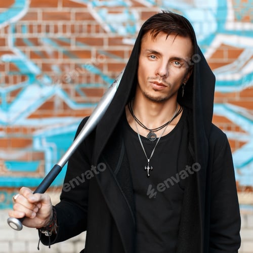 Preview: Handsome man with a bat in black clothes in the hood near the brick wall with graffiti