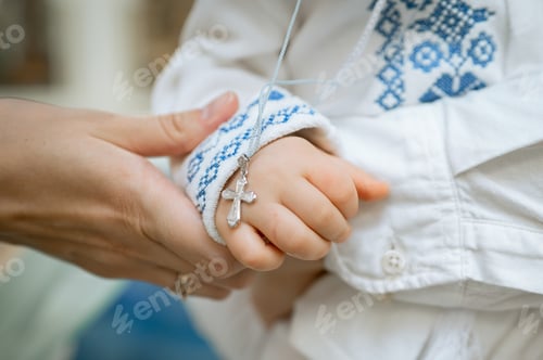 Preview: Baby in traditional embroidered shirt holding silver orthodox cross during baptism ceremony