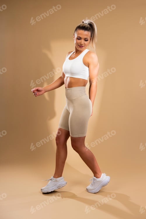 Preview: Fit young woman in white sports bra and beige leggings walking confidently in studio setting