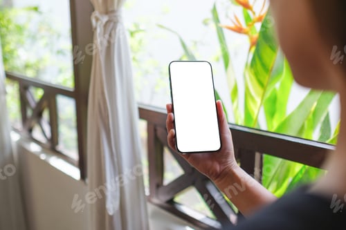Preview: Mockup image of a woman holding mobile phone with blank white desktop screen in tropical style home