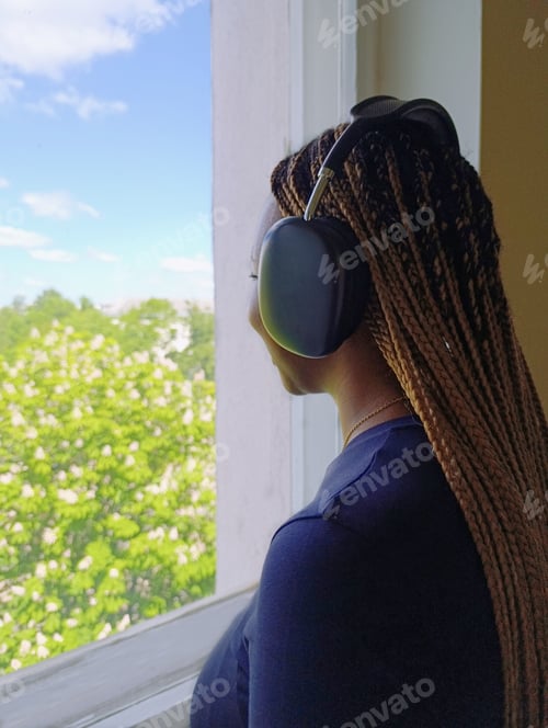 Preview: A young African-American woman with headphones on is standing by the window, listening to music and