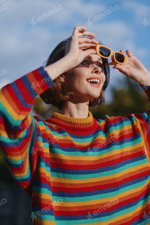 Preview: Woman Wearing Colorful Striped Sweater and Yellow Sunglasses Smiling and Looking up Outdoors