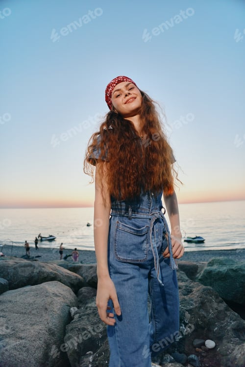 Preview: Smiling Woman With Red Bandana Wearing Denim Dress Standing on Rocky Beach at Sunset With Calm Sea