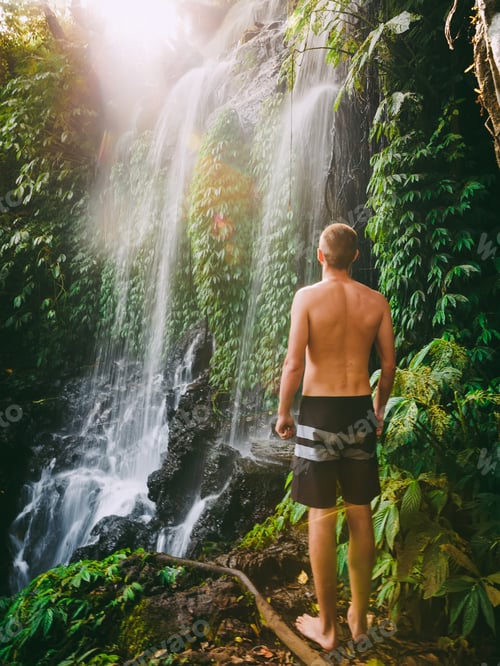 Preview: Male traveler standing beside tropical waterfall in lush jungle landscape